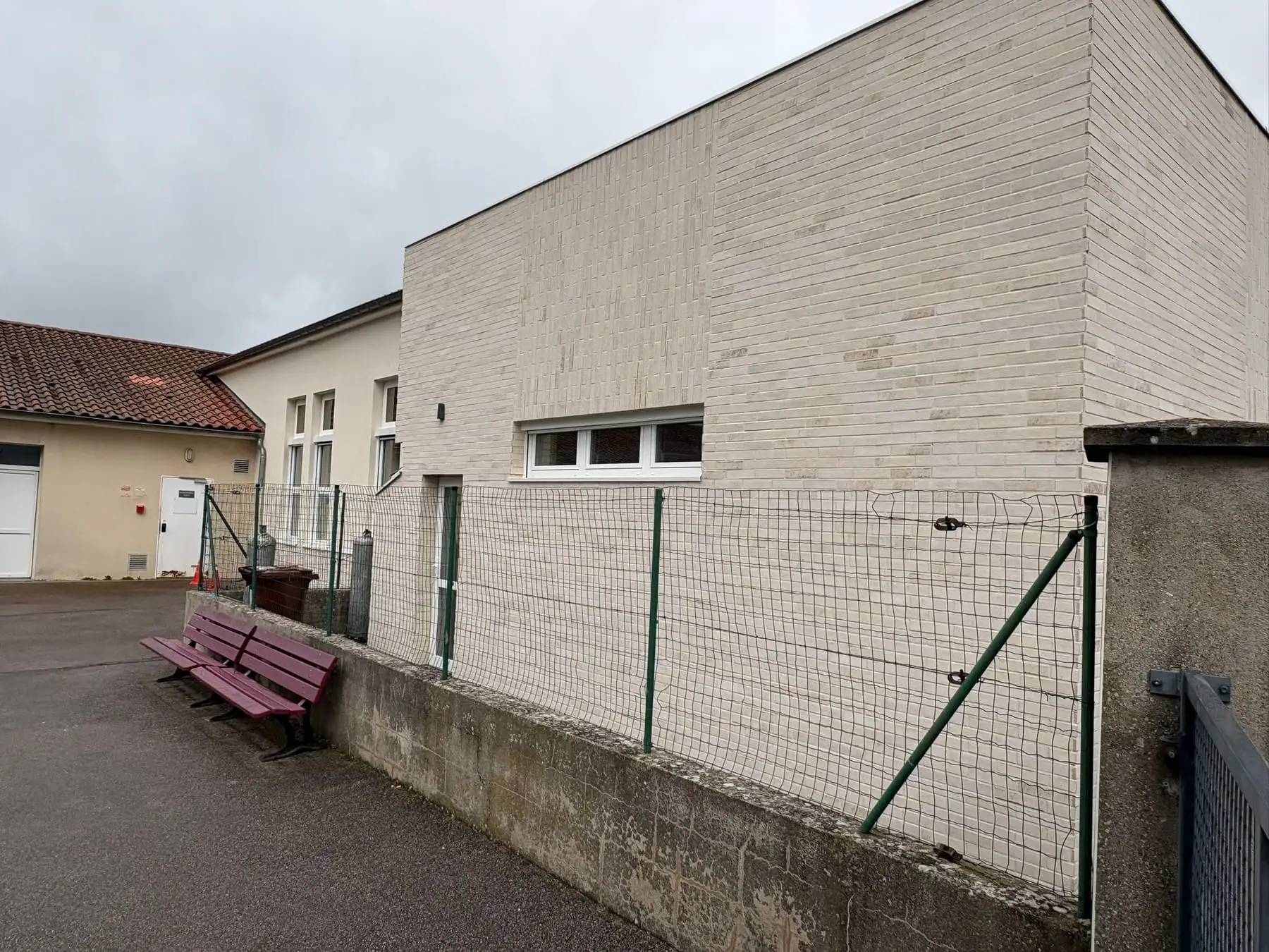 Pignon aveugle habillé de plaquettes de parement claires, avec une fenêtre bandeau et un ancien bâtiment à toiture tuile à gauche.