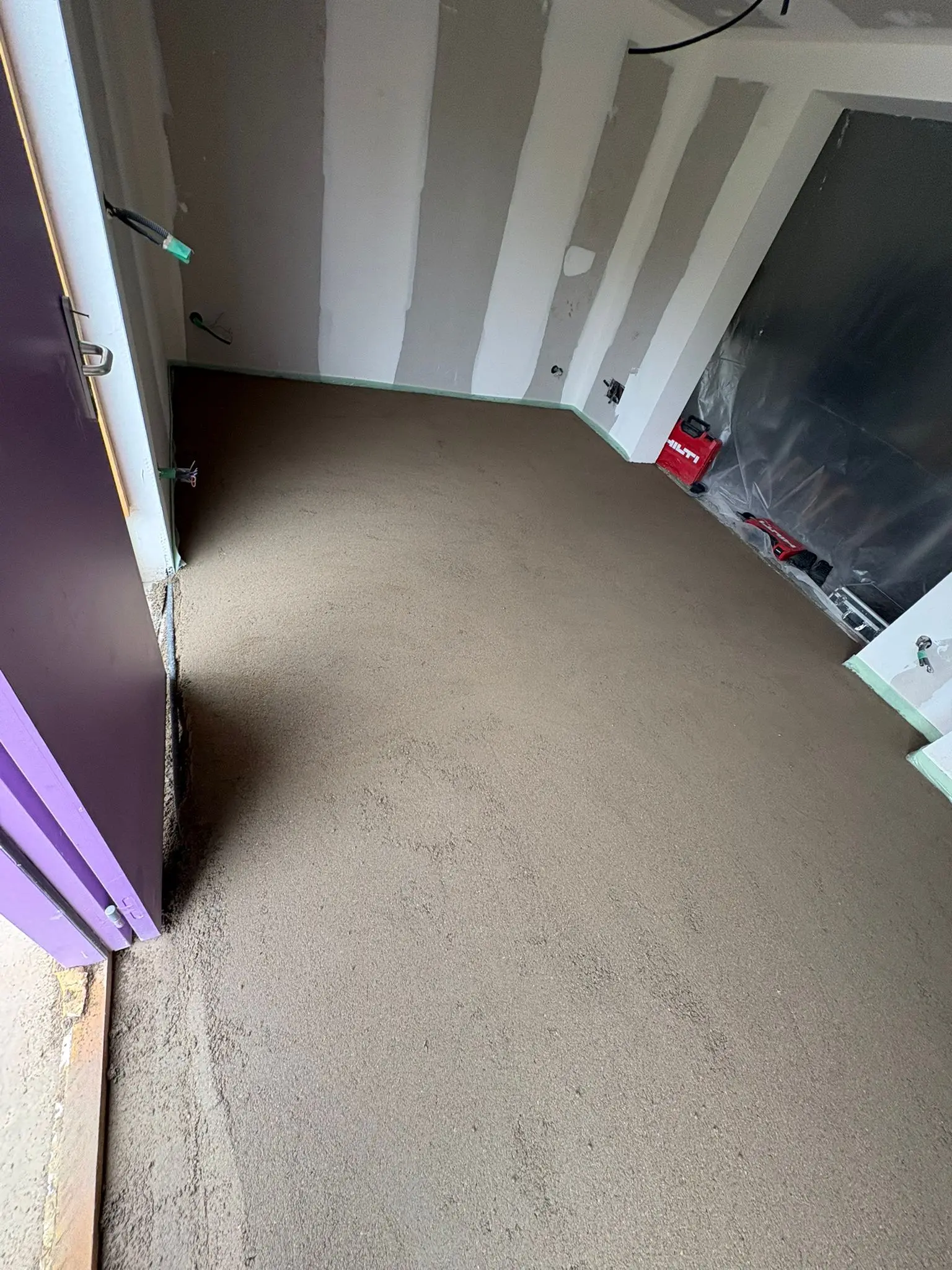 Chape traditionnelle fraîchement tirée dans une pièce doublée en plaques de plâtre, porte violette à gauche et matériel de chantier au fond.