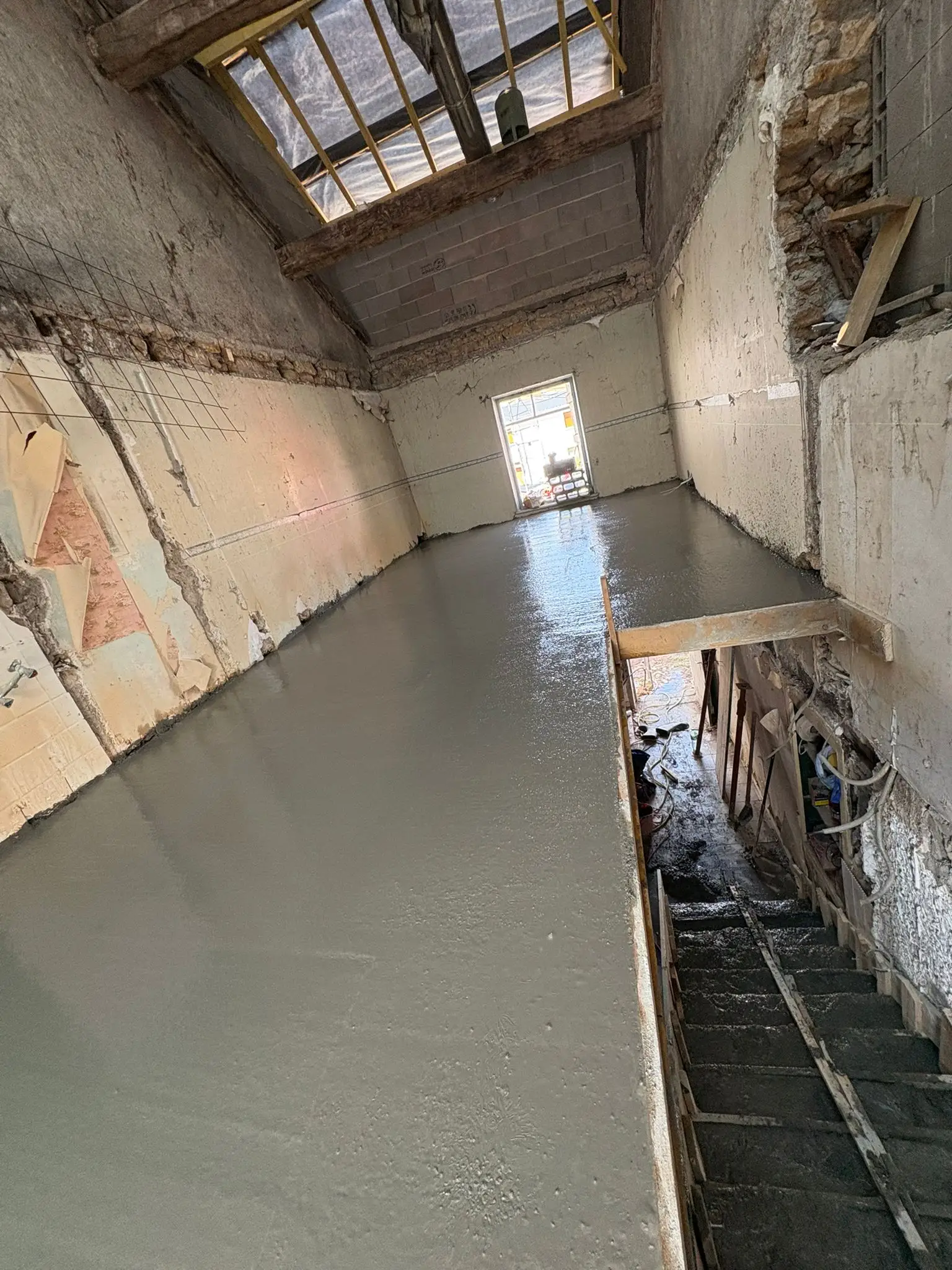 Dalle béton fraîchement coulée et lissée dans une grande pièce en rénovation, murs décroûtés et trémie d’escalier ouverte à droite.