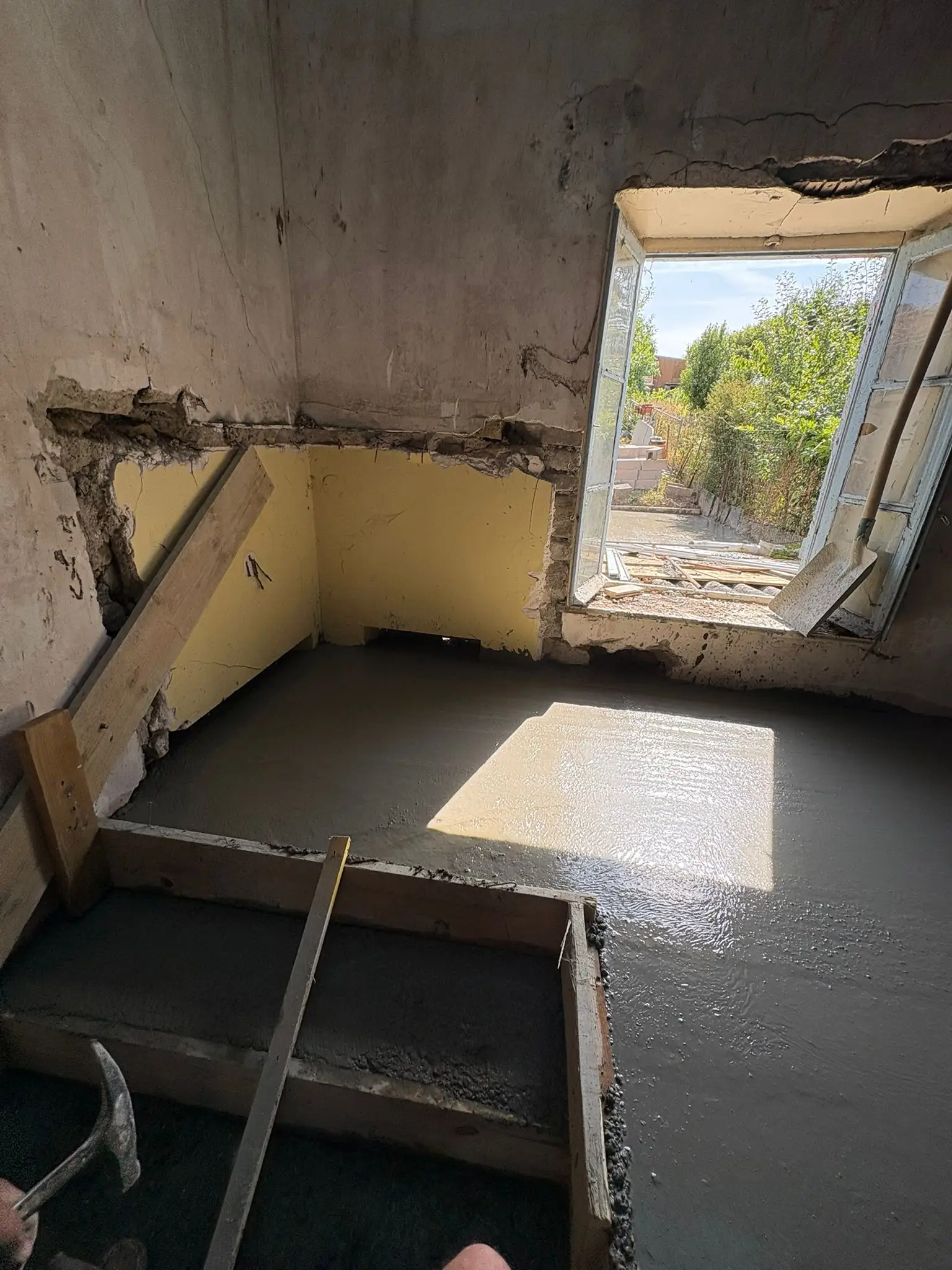 Dalle béton fraîche dans une petite pièce aux murs jaunes écaillés, coffrage bois en rive et vieille fenêtre ouverte.