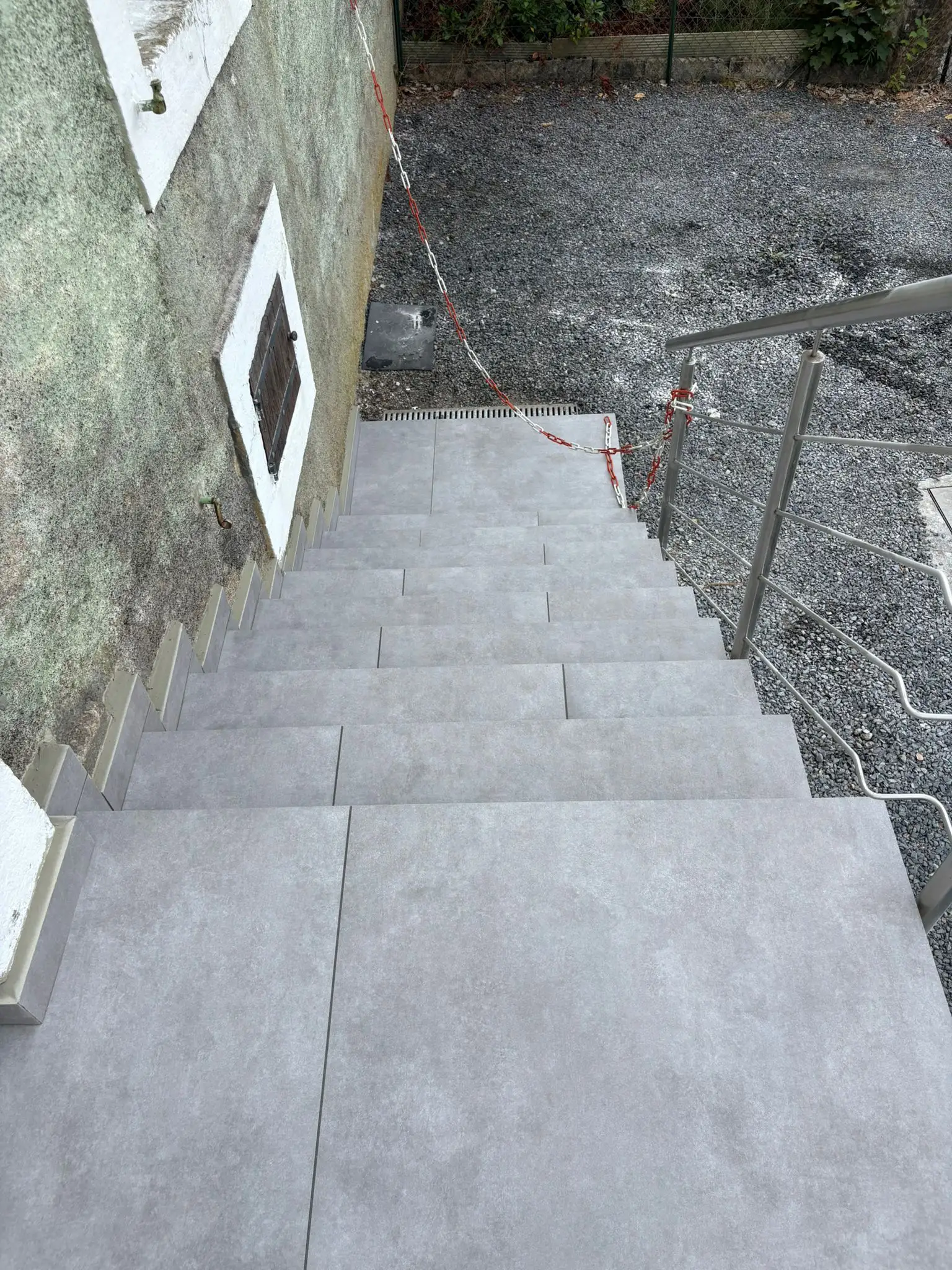 Escalier extérieur vu du haut, marches et palier habillés de dalles grand format gris clair, garde-corps inox à droite.