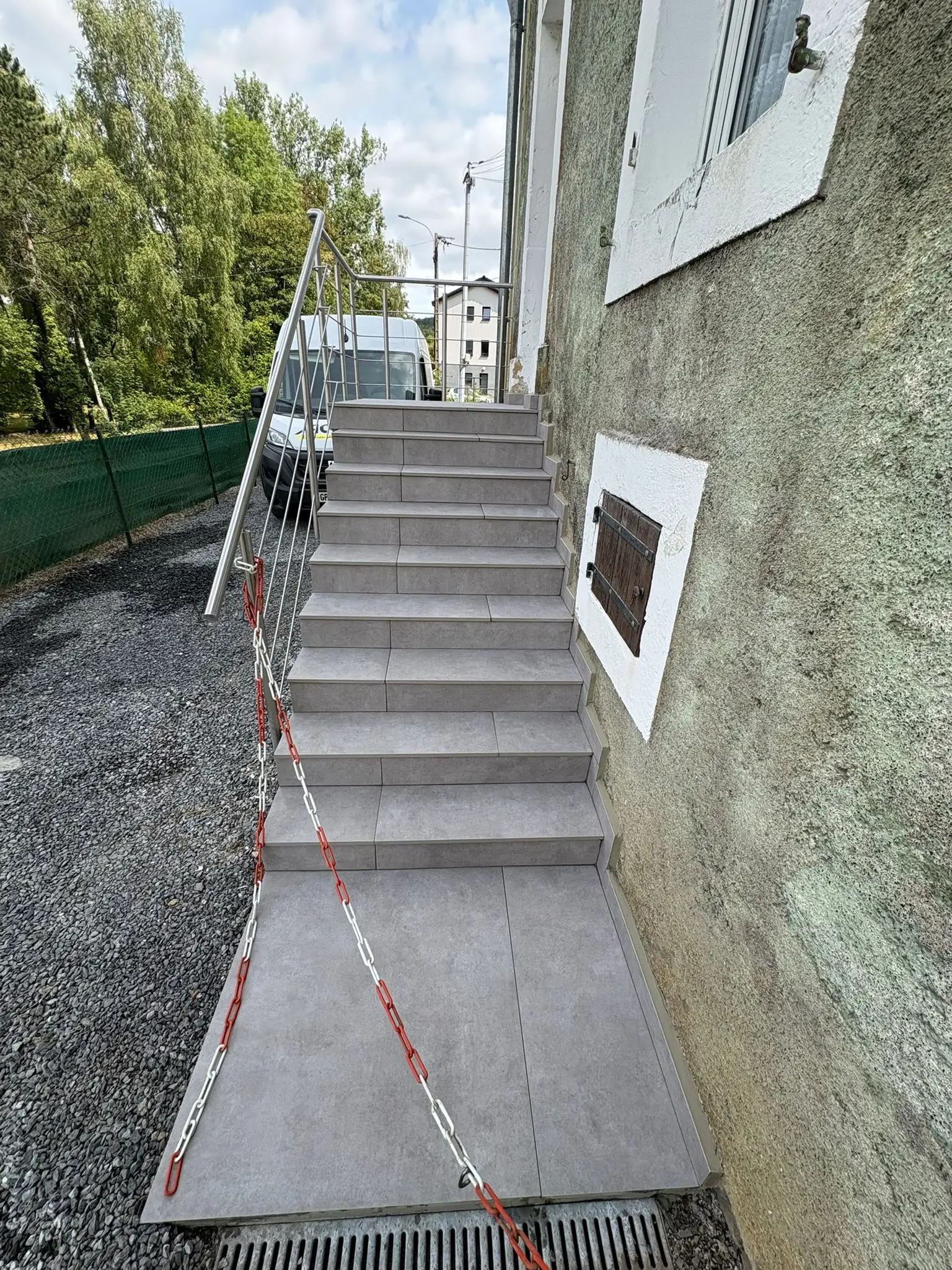 Escalier extérieur et palier habillés de dalles grand format gris, vus depuis le haut des marches, devant une maison enduite.