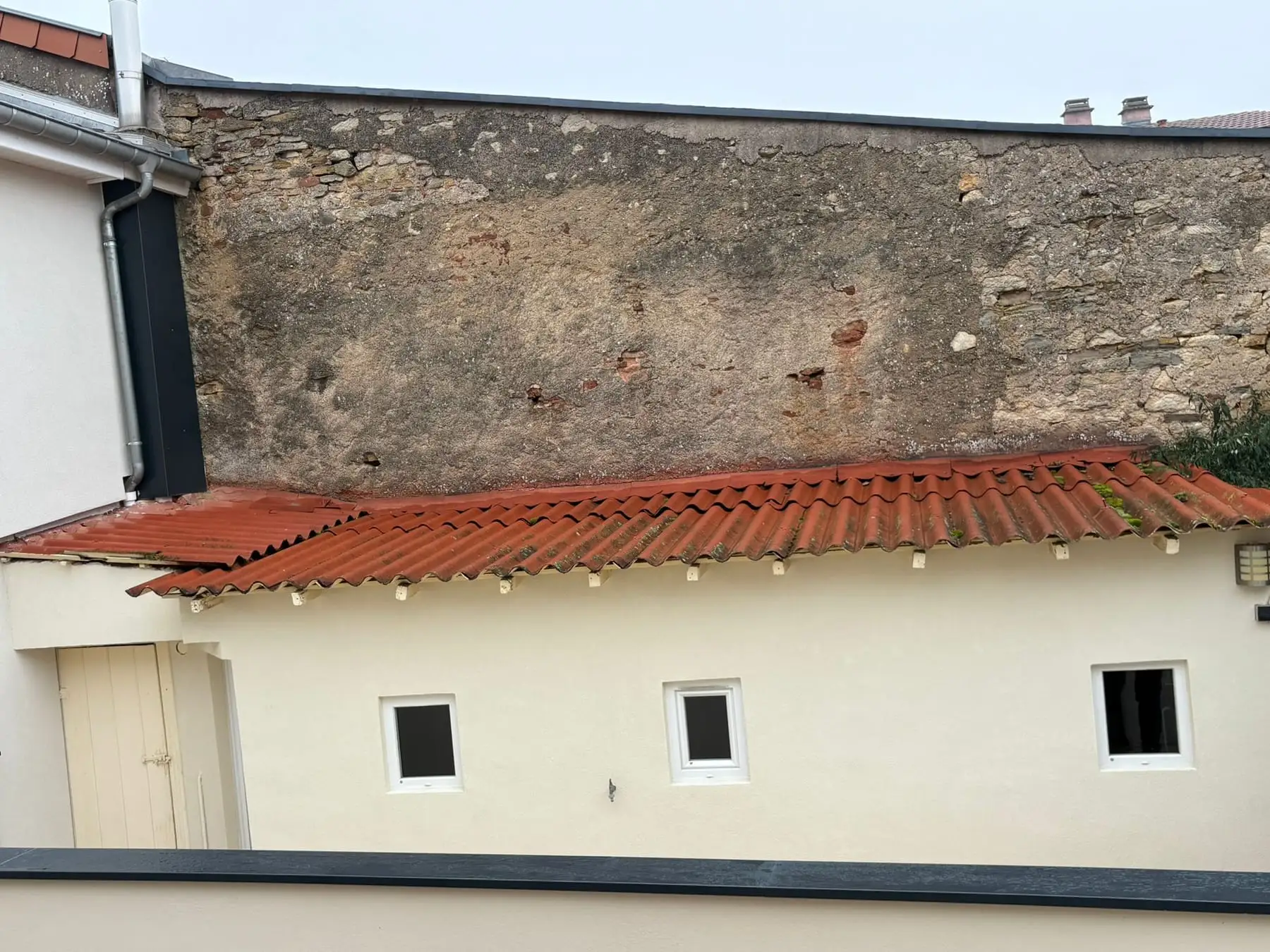 Annexe enduite crème couverte de plaques ondulées rouges, adossée à un haut mur en pierre et enduit ancien.