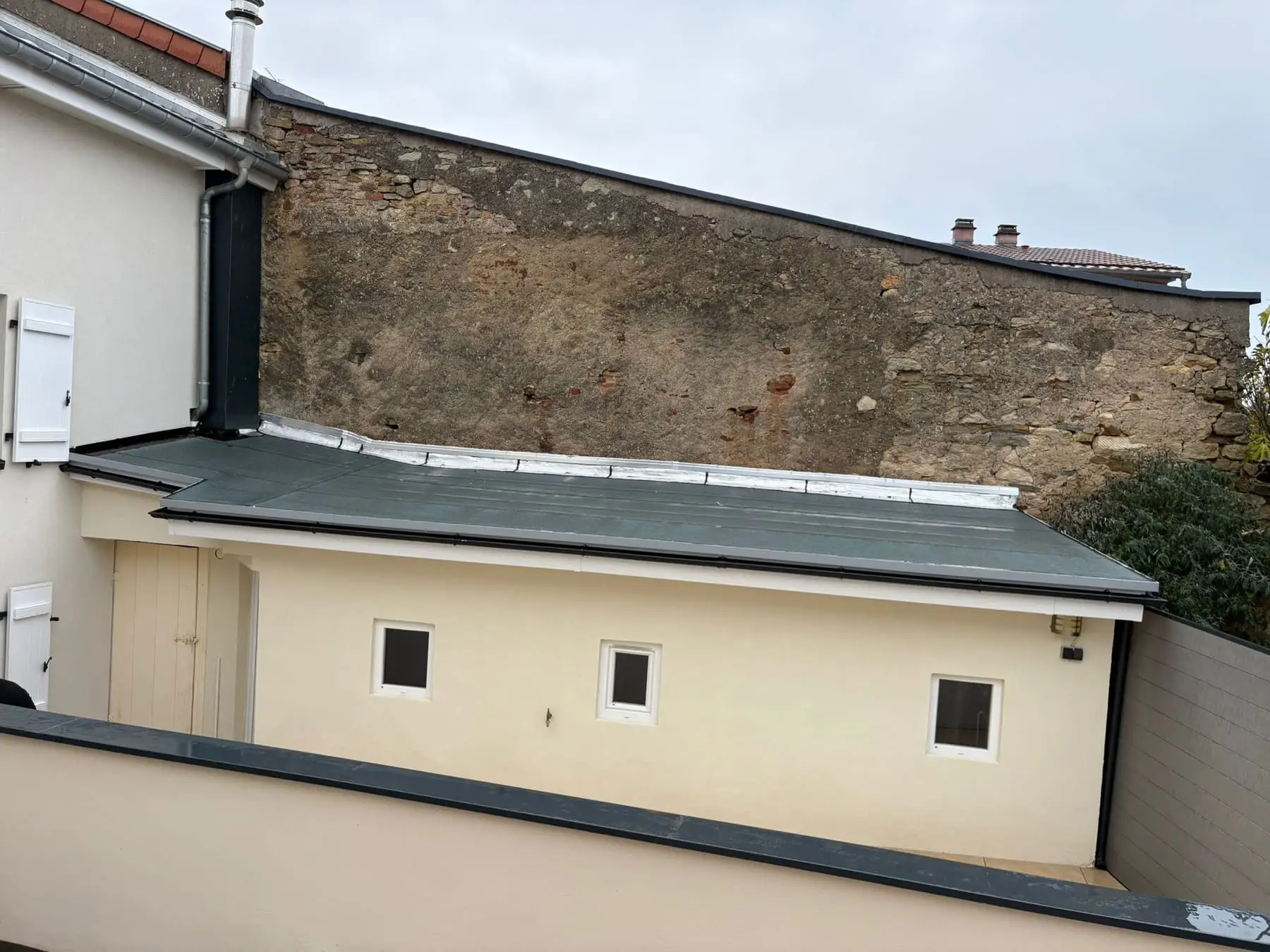 Toiture plate terminée sur une annexe enduite crème, relevé d’étanchéité et bande métallique en tête contre un haut mur en pierre.