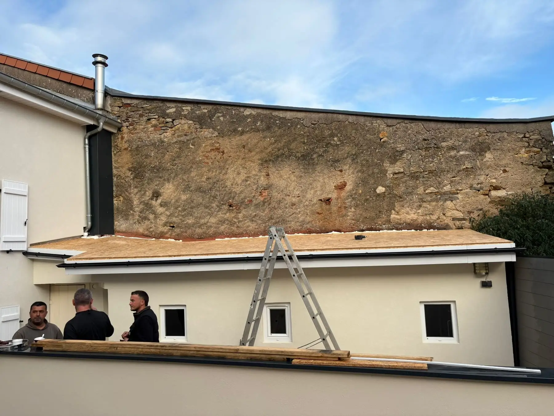 Toiture plate en cours de réalisation, panneaux de bois posés sur la structure, échelle appuyée et compagnons au pied du mur en pierre.