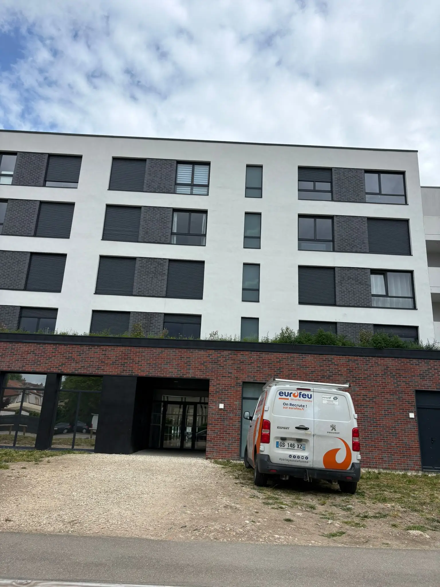 Façade blanche d’un immeuble collectif avec panneaux de briques grises sous les fenêtres et soubassement en briques rouges autour de l’entrée.