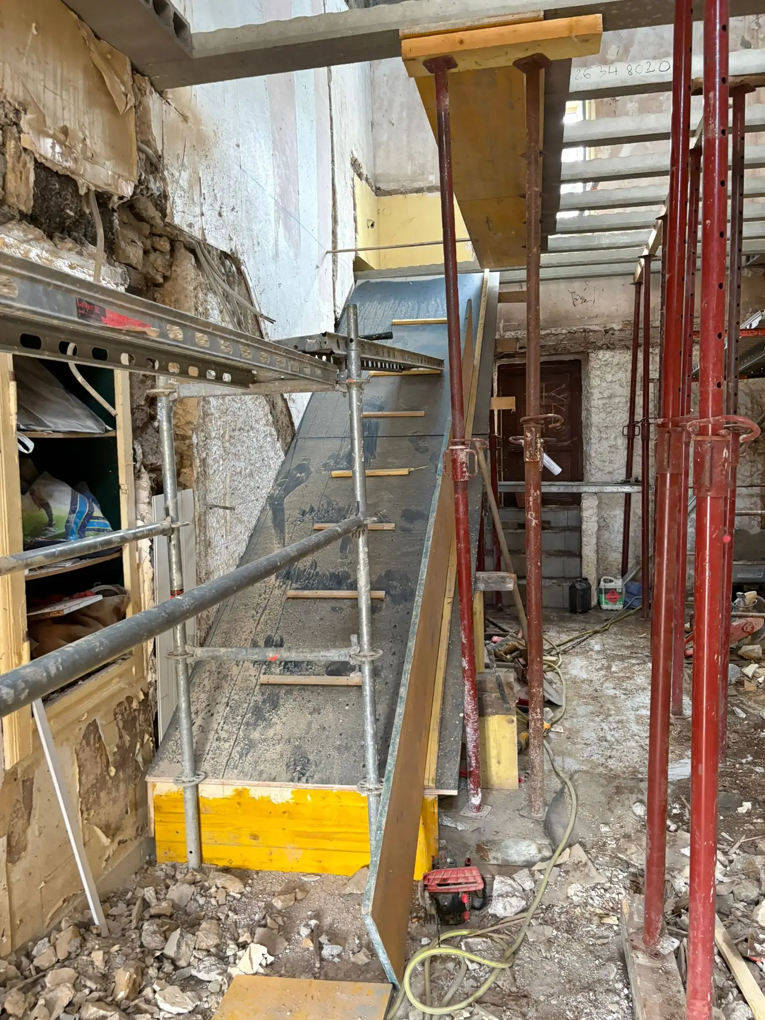 Intérieur en rénovation lourde avec étais métalliques rouges, échafaudage et coffrage d’un escalier béton contre un mur en pierre décroûté.